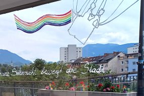 Schaufenstergestaltung bei schau.Räume Villach. Heißluftballonkorb mit Menschen. Regenbogenfahne. Schriftzug: Unsere Sehnsucht bewegt die Stadt.