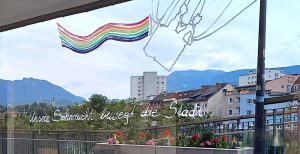 Schaufenstergestaltung bei schau.Räume Villach. Heißluftballonkorb mit Menschen. Regenbogenfahne. Schriftzug: Unsere Sehnsucht bewegt die Stadt.